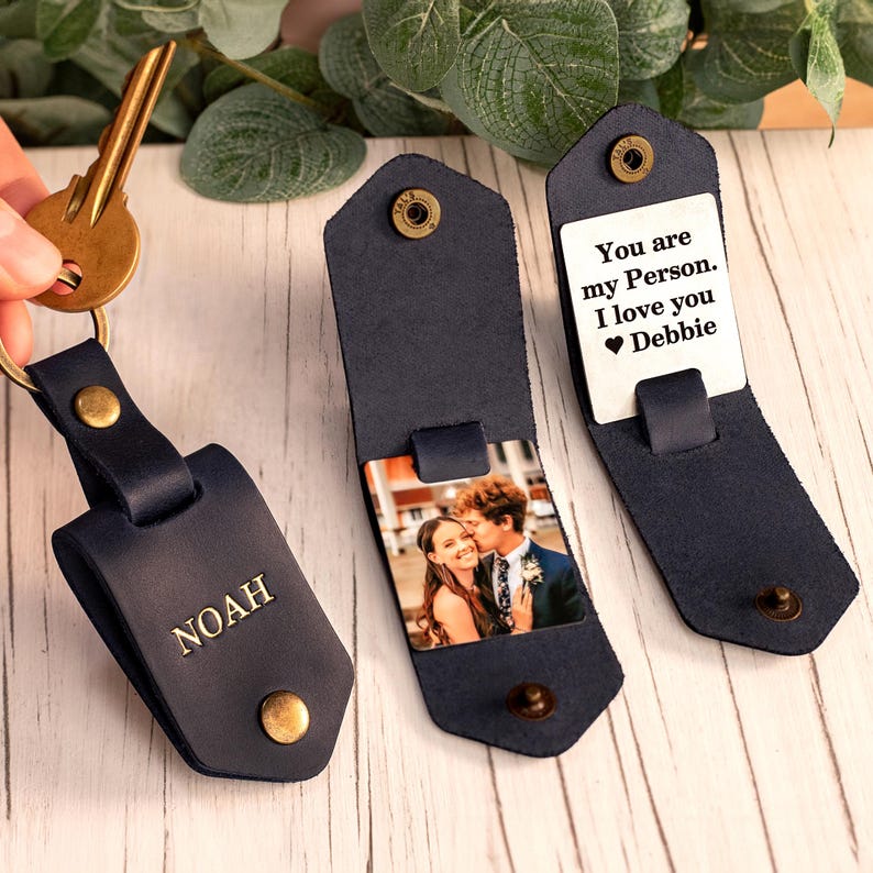 May include: A dark blue leather key organizer with a brass snap closure. One piece is embossed with the name NOAH. Another piece opens to reveal a square photo of a couple and a white metal plate with the text You are my Person. I love you Debbie.