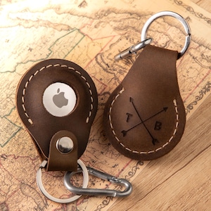 May include: Two dark brown leather keychains with silver metal hardware. One keychain holds a white Apple AirTag, while the other features a stamped design of two crossed arrows with the letters T and B. Both have visible white stitching.