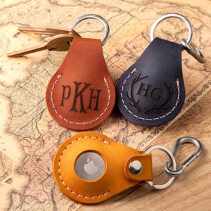 May include: Three leather keyrings with different designs. One is brown with the initials PKH, one is blue with a stag antler design and the initials HG, and one is yellow with a cut-out for an Apple AirTag.