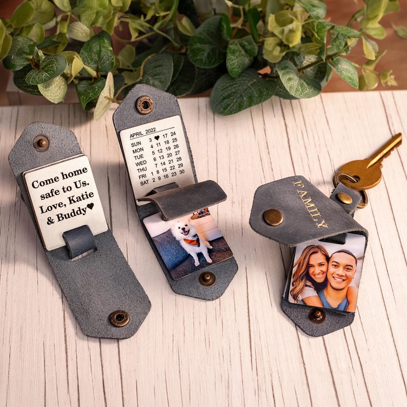 May include: Three grey leather keychains with brass snap closures. One features the text 'Come home safe to us. Love, Katie & Buddy', another shows an April 2022 calendar and a dog photo, and the third displays a photo of two people and the word 'FAMILY'.