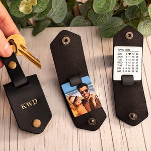 May include: Three black leather key organizers with brass snap buttons. One features the gold embossed letters KWD, another displays a square color photograph of a couple on a beach, and the third shows a white April 2022 calendar card.