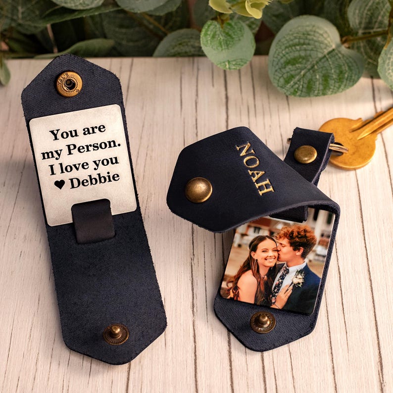 May include: Two dark blue leather key organizers with brass snap buttons. One shows a metal plate engraved with "You are my Person. I love you Debbie". The other is folded, embossed with "NOAH", and displays a photo of a couple inside.