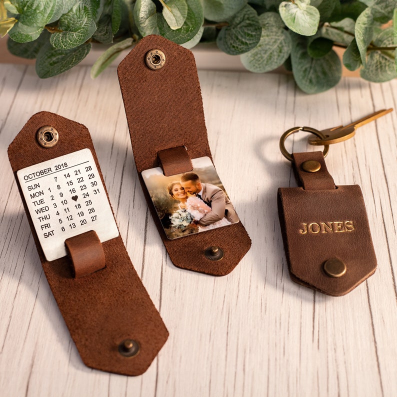 May include: Three brown leather keychains with brass snap closures. One keychain displays a calendar for October 2018 with a heart on the 16th. Another features a square photo of a couple. The third is closed and embossed with the name JONES.