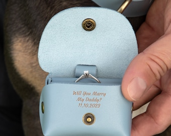 May include: A light blue leatherette ring box with a silver engagement ring inside. The box is open, revealing the ring and the engraved text "Will You Marry My Daddy? 11.10.2023". The box is secured with a snap closure.