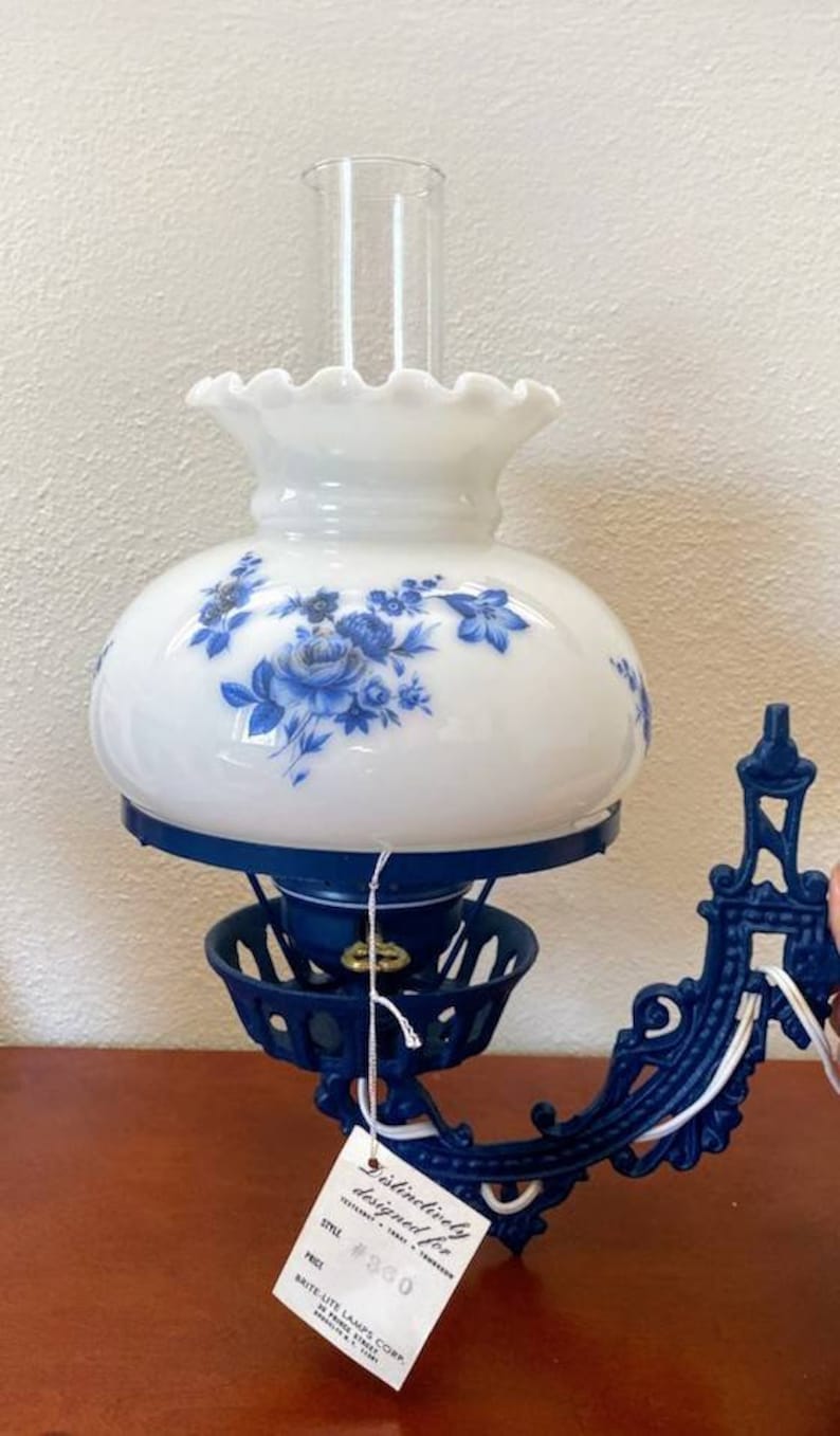 May include: A blue and white oil lamp with a floral design on the glass shade. The lamp is attached to a blue metal wall mount. The lamp has a price tag that reads "Distinctively designed for... #360".