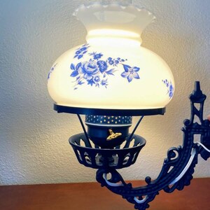 May include: A black cast iron wall sconce with a white glass lampshade featuring a blue floral design. The lampshade has a scalloped edge and a clear glass chimney.