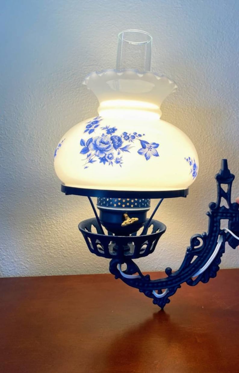 May include: A black cast iron wall sconce with a white glass lampshade featuring a blue floral design. The lampshade has a scalloped edge and a clear glass chimney.