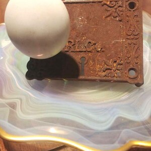 May include: A vintage door knob and lock plate with ornate details. The knob is white porcelain and the plate is rusty metal with a keyhole.