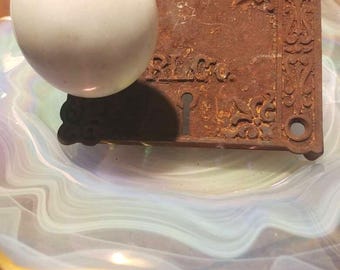 May include: A vintage door knob and lock plate with ornate details. The knob is white porcelain and the plate is rusty metal with a keyhole.