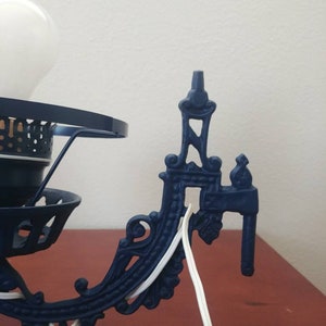 May include: A blue metal wall sconce with ornate detailing and a white electrical cord. The sconce has a round, open-top design with a white light bulb.