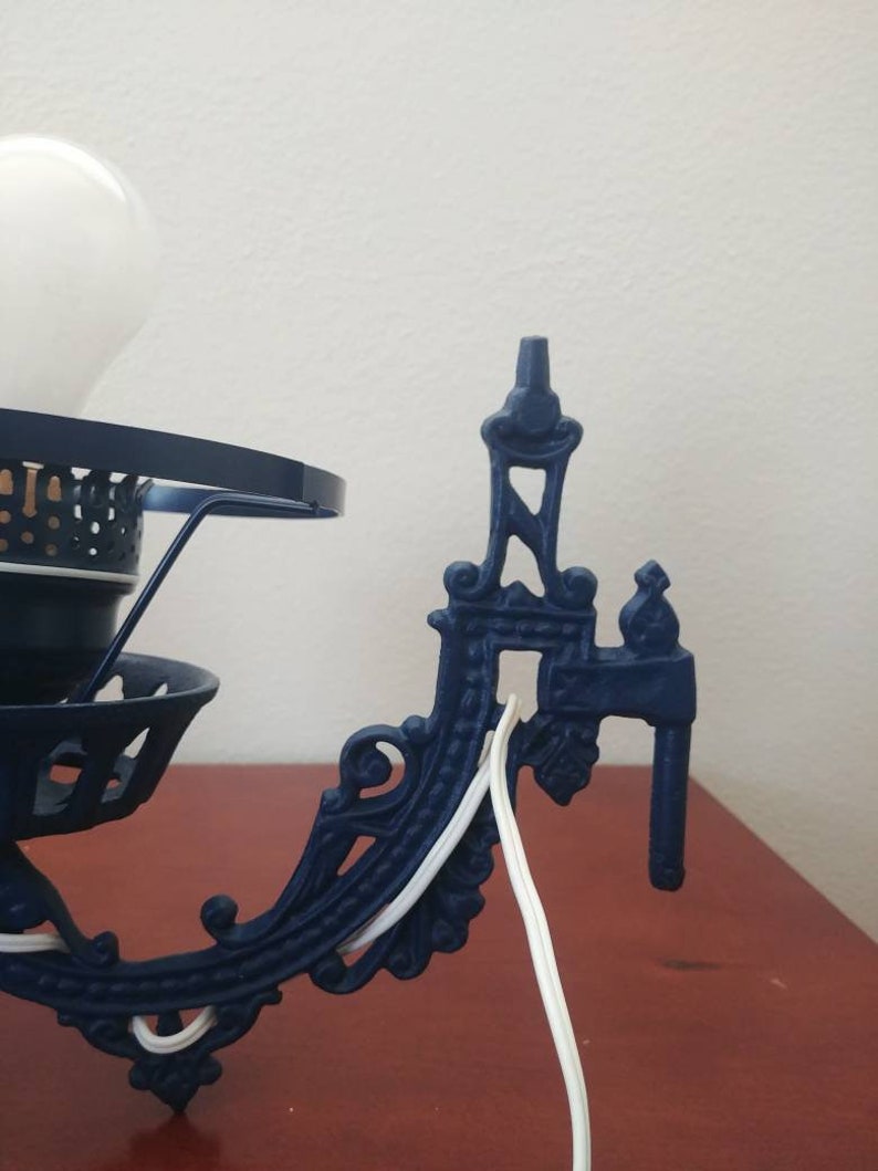 May include: A blue metal wall sconce with ornate detailing and a white electrical cord. The sconce has a round, open-top design with a white light bulb.