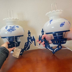 May include: Pair of blue and white wall-mounted lamp fixtures with white glass shades featuring a blue floral design. The shades are attached to a blue metal base with a decorative scroll design.