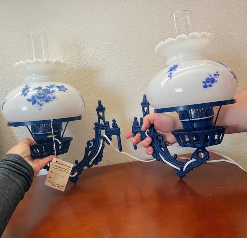 May include: Pair of blue and white wall-mounted lamp fixtures with white glass shades featuring a blue floral design. The shades are attached to a blue metal base with a decorative scroll design.