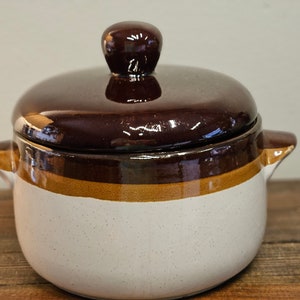 May include: A brown and white ceramic crock pot with a lid. The crock pot has a brown rim and a brown band around the middle. The lid has a brown knob.