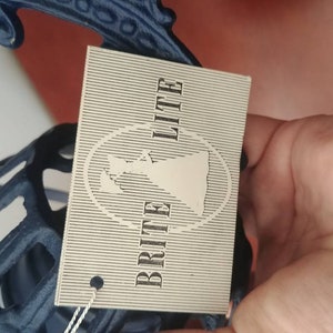 May include: A white tag with black text that reads "Brit Lite" with a stylized image of a woman in a dress. The tag is attached to a blue metal object.