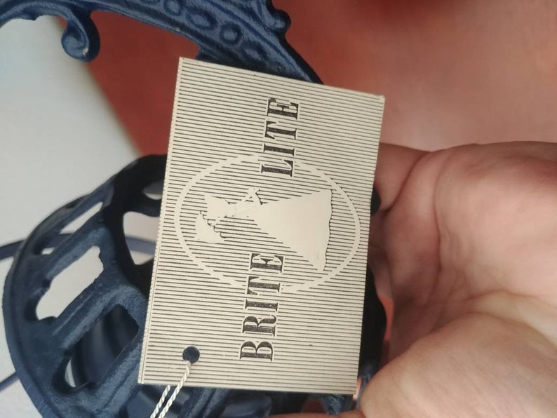 May include: A white tag with black text that reads "Brit Lite" with a stylized image of a woman in a dress. The tag is attached to a blue metal object.