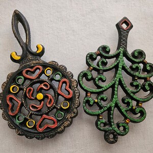 May include: Two black metal trivets with intricate designs. One trivet features a swirling pattern with red, yellow, and green accents. The other trivet has a leafy design with green and yellow accents.