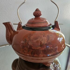 May include: A brown ceramic teapot with a floral design and a spring-loaded handle. The teapot has a black rim and a black stripe around the middle. The teapot is sitting on a reflective surface.