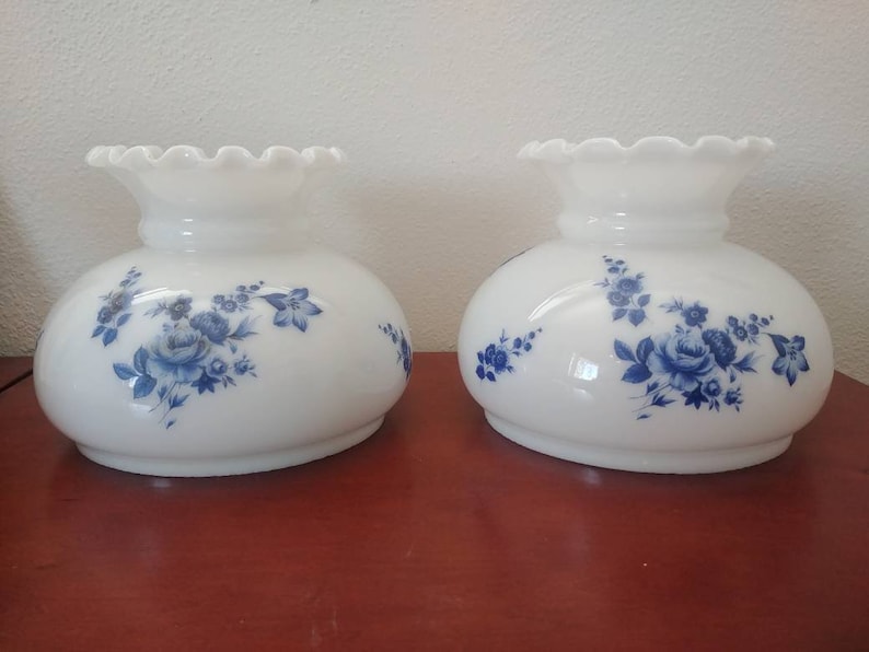 May include: Two white glass lampshades with a blue floral pattern. The lampshades have a scalloped edge.