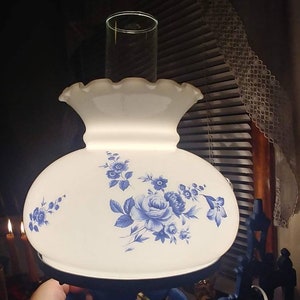 May include: A white and blue floral lampshade with a blue metal base. The lampshade has a ruffled edge and a floral design with blue flowers on a white background. The base is a dark blue metal with a gold accent.