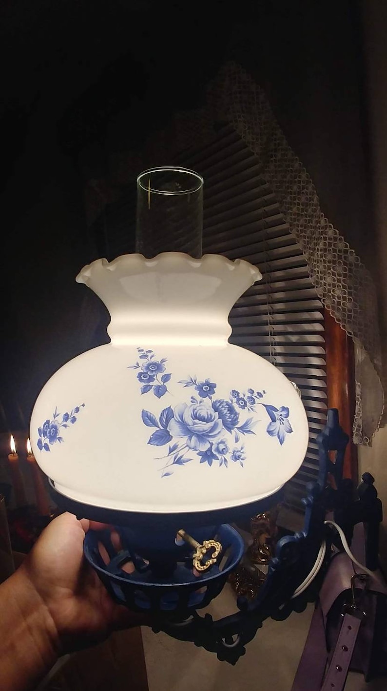 May include: A white and blue floral lampshade with a blue metal base. The lampshade has a ruffled edge and a floral design with blue flowers on a white background. The base is a dark blue metal with a gold accent.
