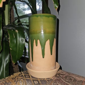 May include: A cylindrical ceramic planter with a green glaze that drips down over a tan base. The planter sits on a matching saucer. The background includes green plants and a window.