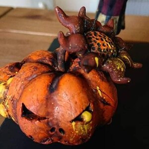 May include: A ceramic pumpkin with a scary face and a brown and yellow spider on top. The pumpkin is painted with orange, yellow, and brown paint.