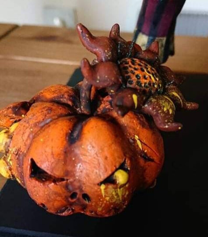 May include: A ceramic pumpkin with a scary face and a brown and yellow spider on top. The pumpkin is painted with orange, yellow, and brown paint.