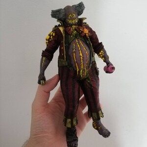 May include: A hand-painted, 3D printed figurine of a clown with a grotesque, yellow and black face, wearing a red and gold costume with a yellow and black pattern. The clown is holding a pink object in its right hand.