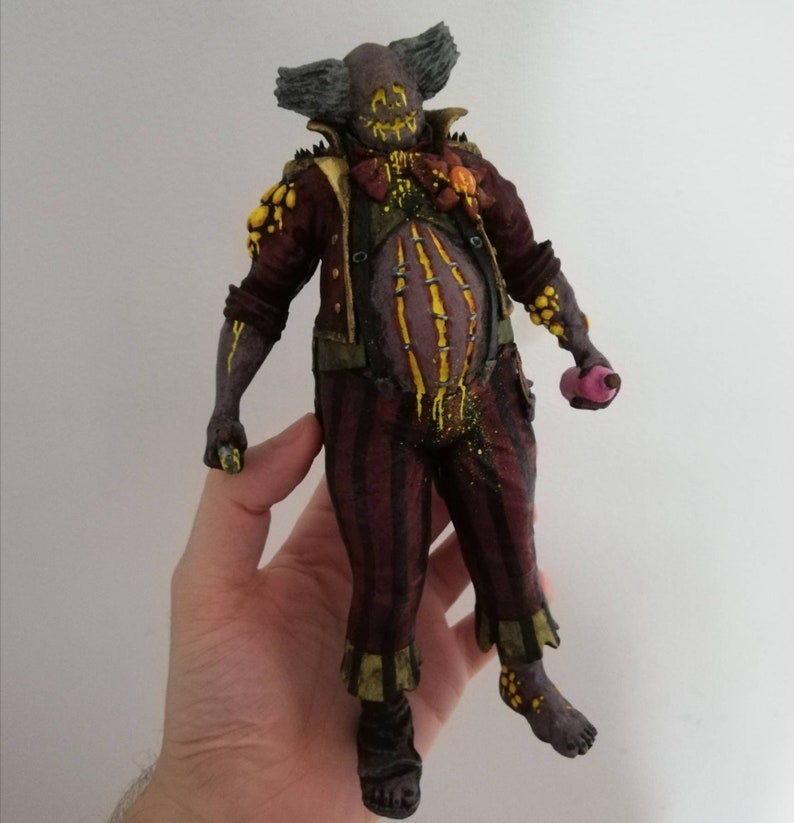 May include: A hand-painted, 3D printed figurine of a clown with a grotesque, yellow and black face, wearing a red and gold costume with a yellow and black pattern. The clown is holding a pink object in its right hand.