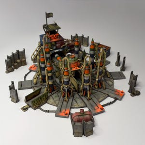 May include: A detailed miniature model of a military installation, featuring multiple missile launch platforms, a control tower, and various support structures. The model is painted in shades of gray, orange, and green, with a weathered appearance. A flag is atop the control tower.