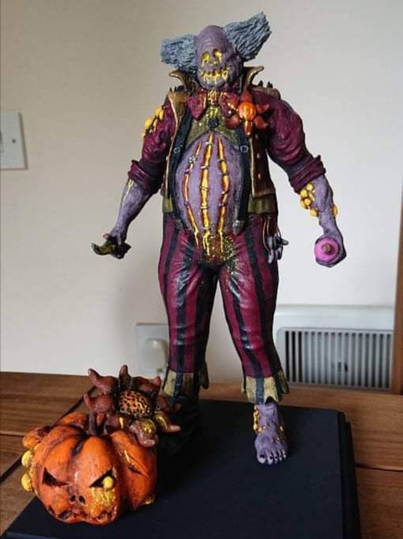 May include: A creepy clown figurine with a purple and red costume, a large belly, and a pumpkin with a scary face. The clown is holding a pink donut in one hand and a small object in the other.