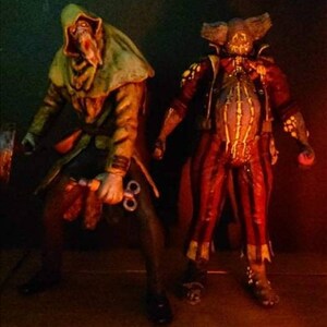 May include: Two horror-themed action figures. The figure on the left is wearing a green hooded cloak and holding a pair of scissors. The figure on the right is wearing a red and yellow jester costume with a stitched-up face.