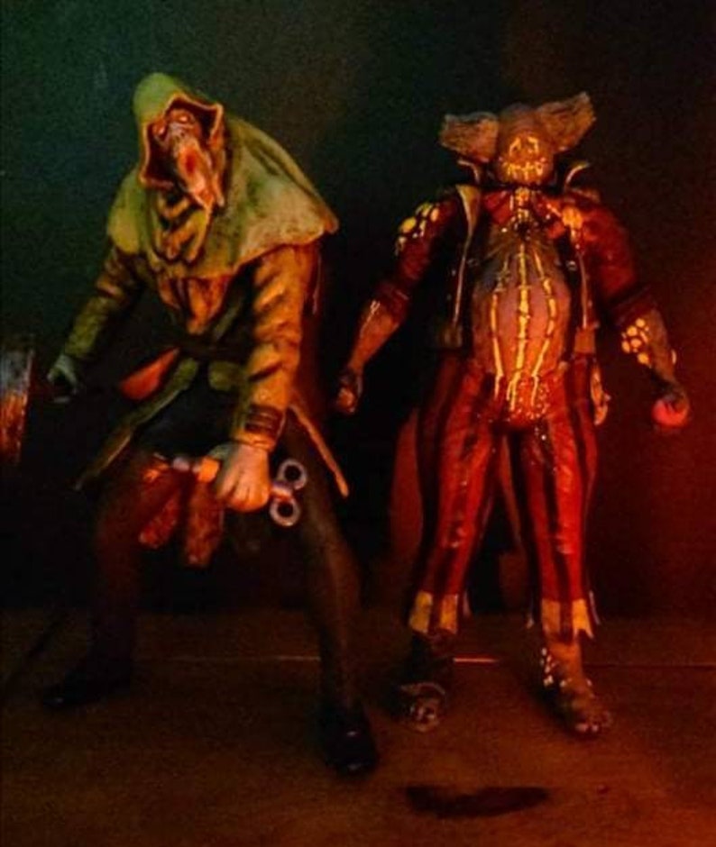 May include: Two horror-themed action figures. The figure on the left is wearing a green hooded cloak and holding a pair of scissors. The figure on the right is wearing a red and yellow jester costume with a stitched-up face.