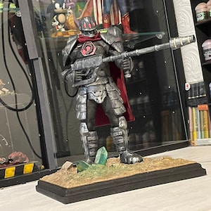 May include: A detailed, hand-painted action figure of a futuristic soldier wearing silver armour and a red cape. The figure is holding a large, silver gun and is standing on a base with green and brown accents.