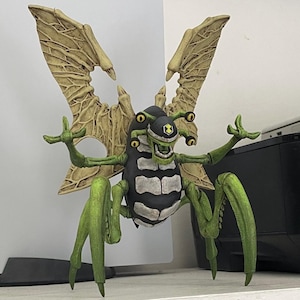 May include: A green and black insect-like figure with large, skeletal wings. The figure has multiple eyes and a mouth with sharp teeth. The figure is standing on a white surface.