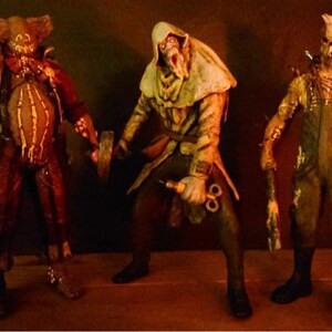 May include: Three horror movie character action figures. The figures are made of plastic and are painted in realistic detail. The figures are posed in a menacing way. The figure on the left is wearing a brown and red costume with a mask. The figure in the middle is wearing a white hooded robe and holding a syringe. The figure on the right is wearing a brown jumpsuit and a mask with a jagged mouth.