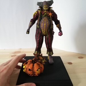 May include: A hand-painted, creepy clown figurine with a pumpkin and candy corn. The clown is wearing a red and black striped outfit with a yellow and brown waistcoat. The pumpkin is orange with a yellow face and black eyes.