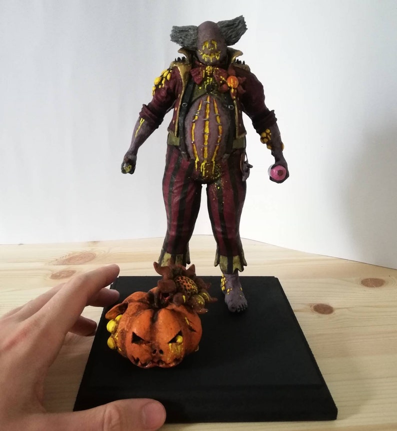 May include: A hand-painted, creepy clown figurine with a pumpkin and candy corn. The clown is wearing a red and black striped outfit with a yellow and brown waistcoat. The pumpkin is orange with a yellow face and black eyes.