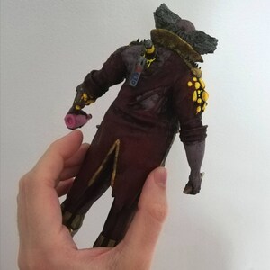 May include: A 3D printed figurine of a character wearing a red and gold costume with yellow accents. The character has a large, bushy beard and is holding a pink object in its hand. The figurine is standing on a white background.