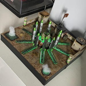 May include: A miniature model of a futuristic military base with a central green structure surrounded by eight glowing green missile launchers. The base is built on a brown, textured surface and includes several small buildings and a flag.