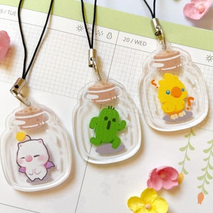 May include: Three clear acrylic charms with black cords. Each charm features a different cartoon character inside a jar: a white creature with purple wings, a green cactus, and a yellow chick. The charms have cork-like tops and are on a planner.