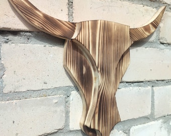 May include: A wooden bull skull wall hanging with a rustic, burnt wood finish. The horns curve outward, and the skull has a layered design. The piece is mounted on a white brick wall, showcasing the natural wood grain.