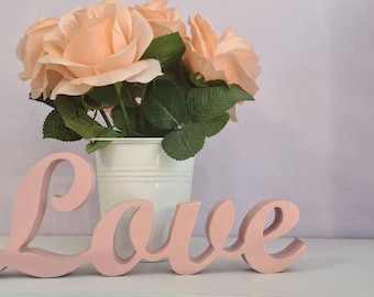 May include: A decorative arrangement featuring a white bucket filled with peach-colored roses and green leaves. The word "Love" is spelled out in a soft pink, wooden script, placed in front of the flowers.
