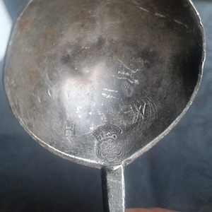 Könnte beinhalten: Nahaufnahme eines angelaufenen Silberlöffels. Der Löffel hat eine abgerundete Schüssel und einen rechteckigen Griff. Die Schüssel hat Markierungen und ein dekoratives Emblem. Der Löffel scheint antik zu sein.