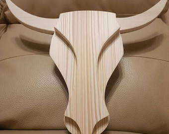 May include: A wooden bull skull wall decor. The sculpture is made of light-colored wood, with visible grain patterns. The bull's horns curve outward, and the design features a detailed, stylized face. This rustic piece is suitable for home or office decor.