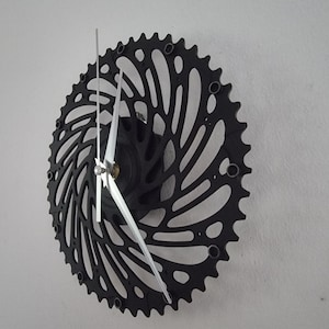 Könnte beinhalten: Schwarze Metalluhr mit einem einzigartigen Design, das einen Fahrradkettenring zeigt. Die Uhrzeiger sind silberfarben und die Uhr ist an einer weißen Wand montiert.