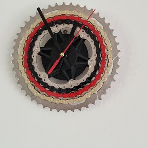 Könnte beinhalten: Eine Wanduhr aus Fahrradkomponenten mit einem großen silbernen Zahnrad, einem goldenen, einem roten und einem schwarzen Kettenblatt. Die Mitte besteht aus einer schwarzen sternförmigen Nabe mit schwarzen Stunden- und Minutenzeigern sowie einem roten Sekundenzeiger.