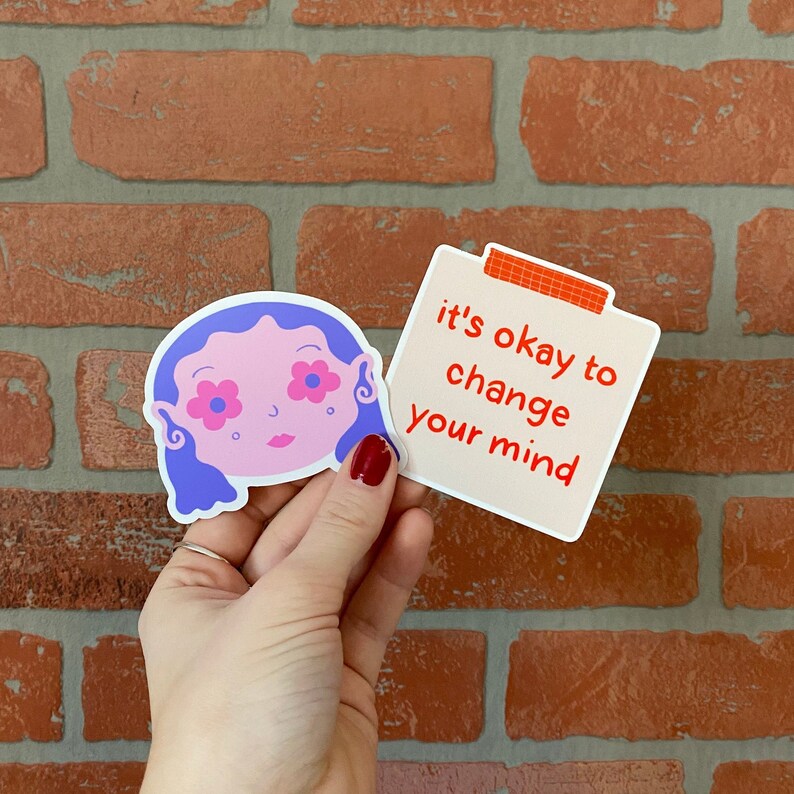 May include: Two stickers, one with a cartoon illustration of a person with blue hair and pink flowers on their eyes, and the other with the text "it's okay to change your mind".
