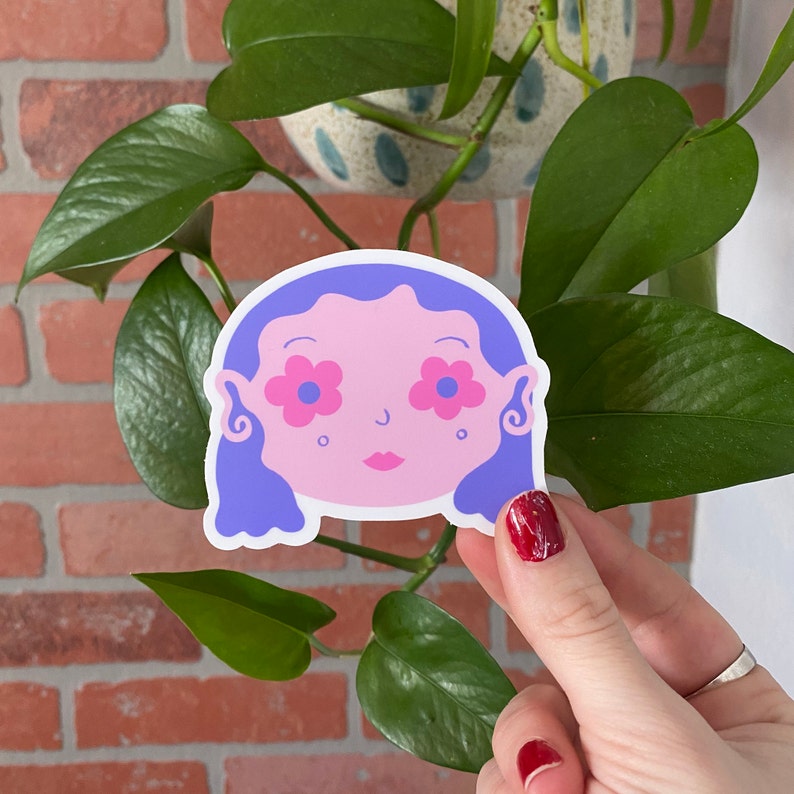 May include: A sticker of a cartoon face with purple hair and pink flowers for eyes. The face is pink and has a small nose and mouth.